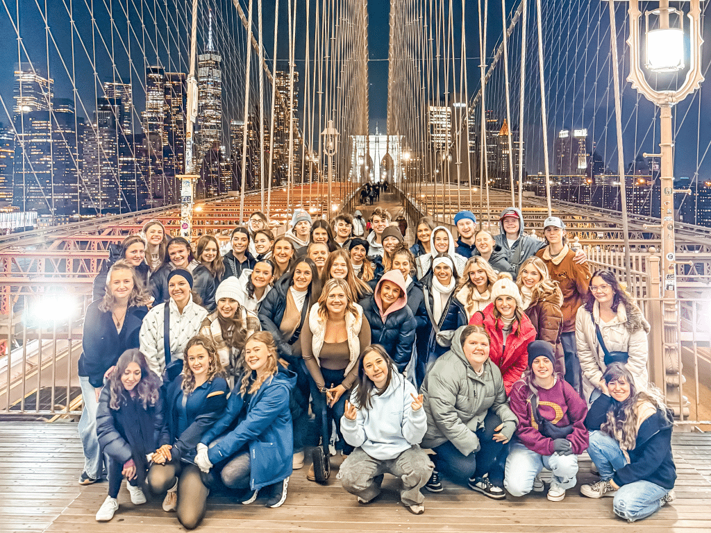 A large group of people poses and smiles for a photo at night on the Brooklyn Bridge during their Landmark Educational Tours Registration, with New York City skyscrapers glowing in the background.