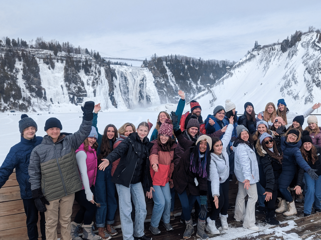 A large group of people in winter clothing poses and smiles in front of frozen waterfalls and snow-covered cliffs under a cloudy sky.