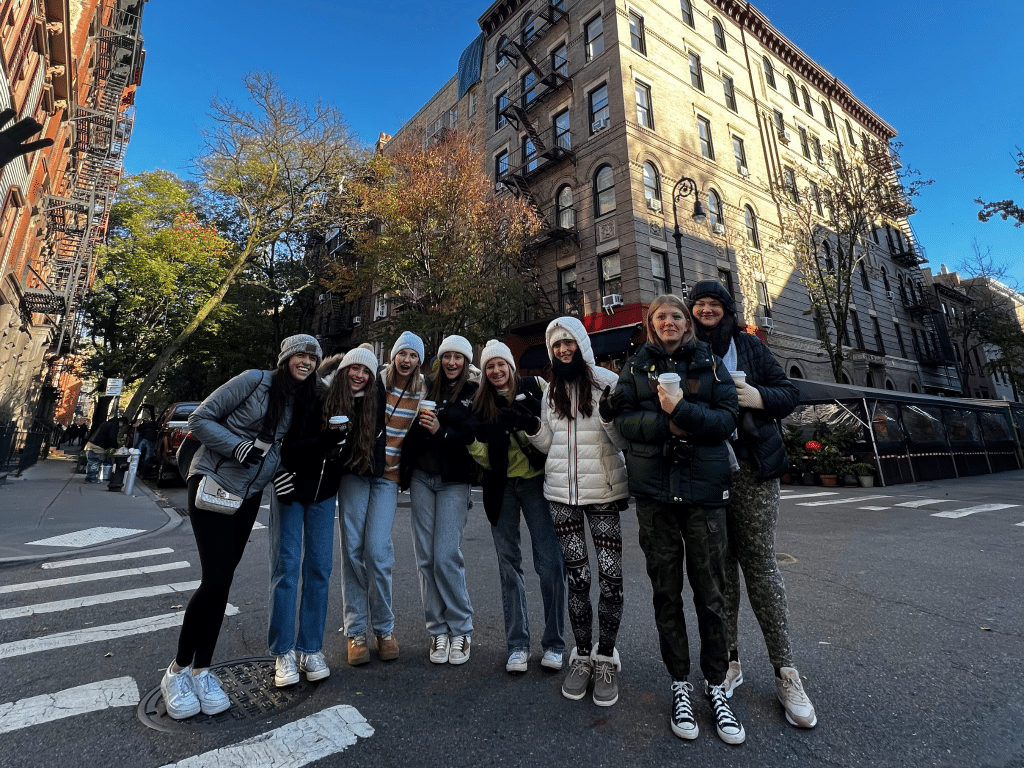Eight people in winter clothes pose together and smile on a city street corner, possibly during Landmark Educational Tours Registration, with tall apartment buildings and trees behind them under a clear blue sky.