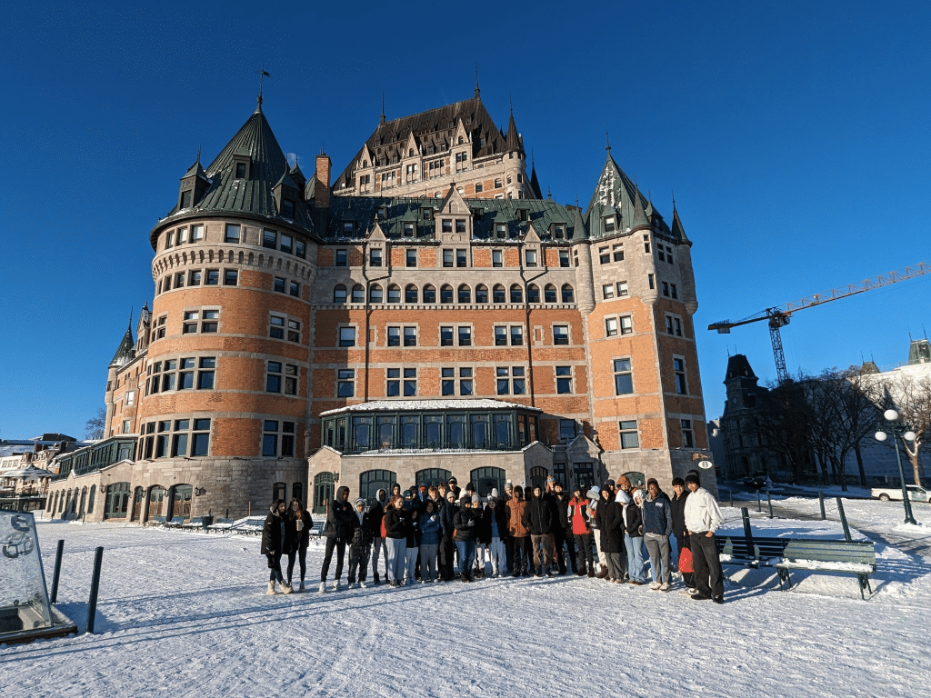 A large group of people stands on snowy ground in front of the historic Château Frontenac, a grand hotel with turrets and steep roofs, under a clear blue sky.