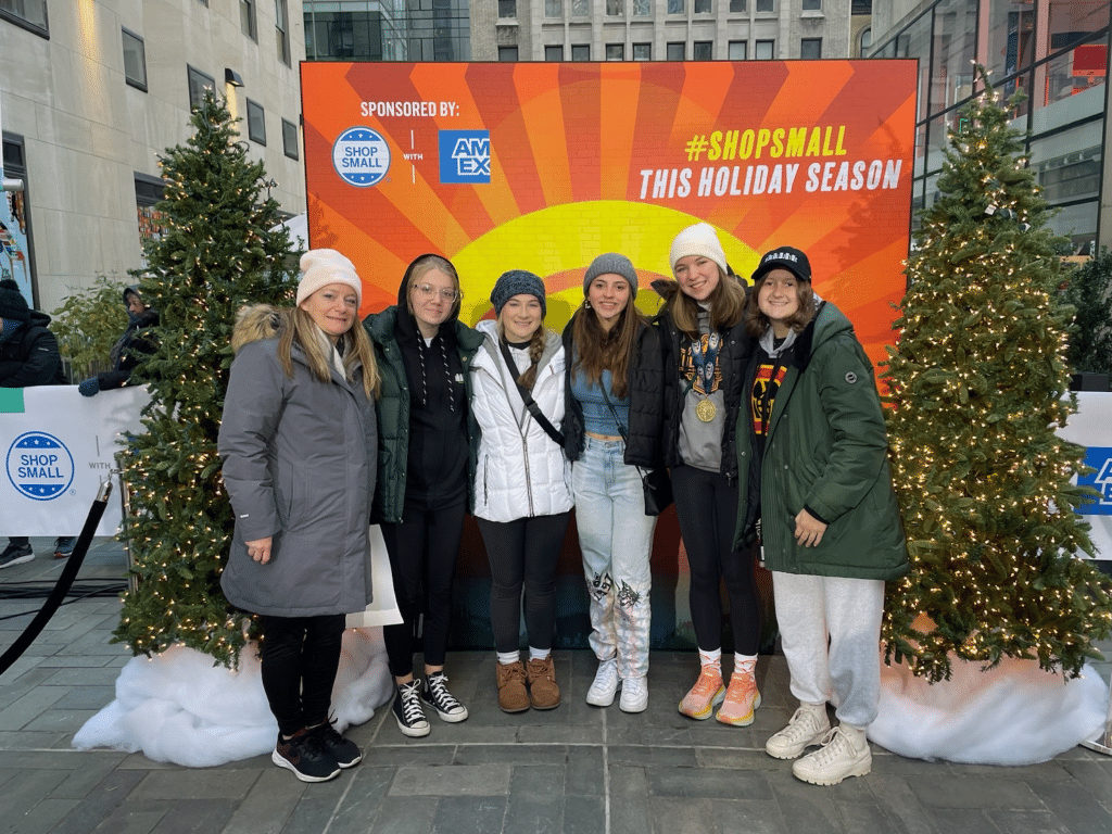 Six young women in winter clothing stand together, smiling in front of a festive holiday backdrop with small decorated Christmas trees and a sign that says, "Shop Small This Holiday Season," "Sponsored by AMEX," and "Landmark Educational Tours Registration.