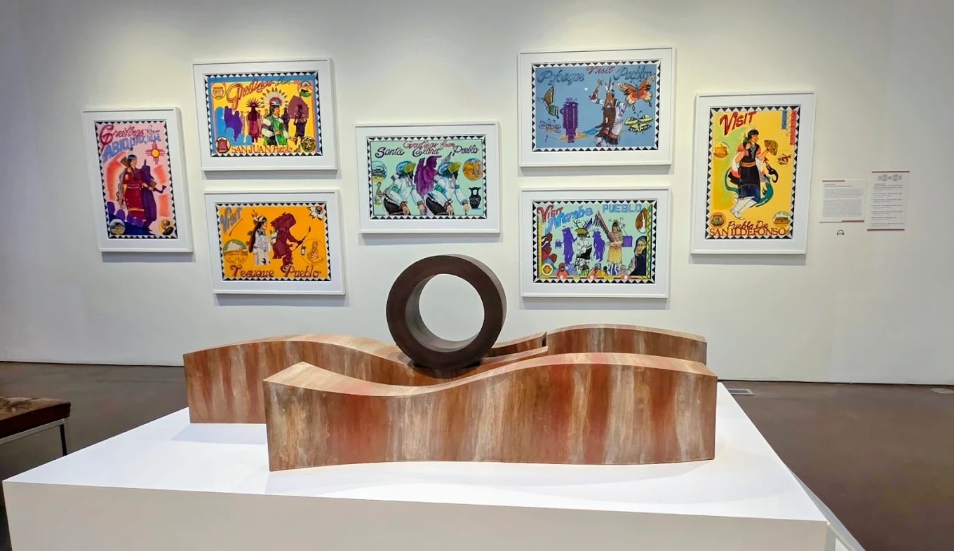 A gallery display featuring a wavy wooden sculpture with a dark circular centerpiece, set in front of a wall with eight colorful, framed artworks depicting vibrant scenes—a perfect Field Trip New Mexico experience for art lovers.
