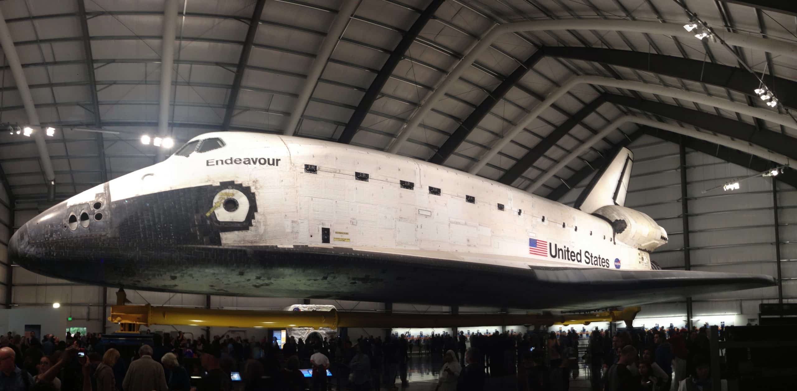 The space shuttle Endeavour is displayed inside a large hangar in Los Angeles, making it a popular destination for Los Angeles field trips. Surrounded by visitors, the white shuttle features black accents, the NASA logo, and "United States" on its side.