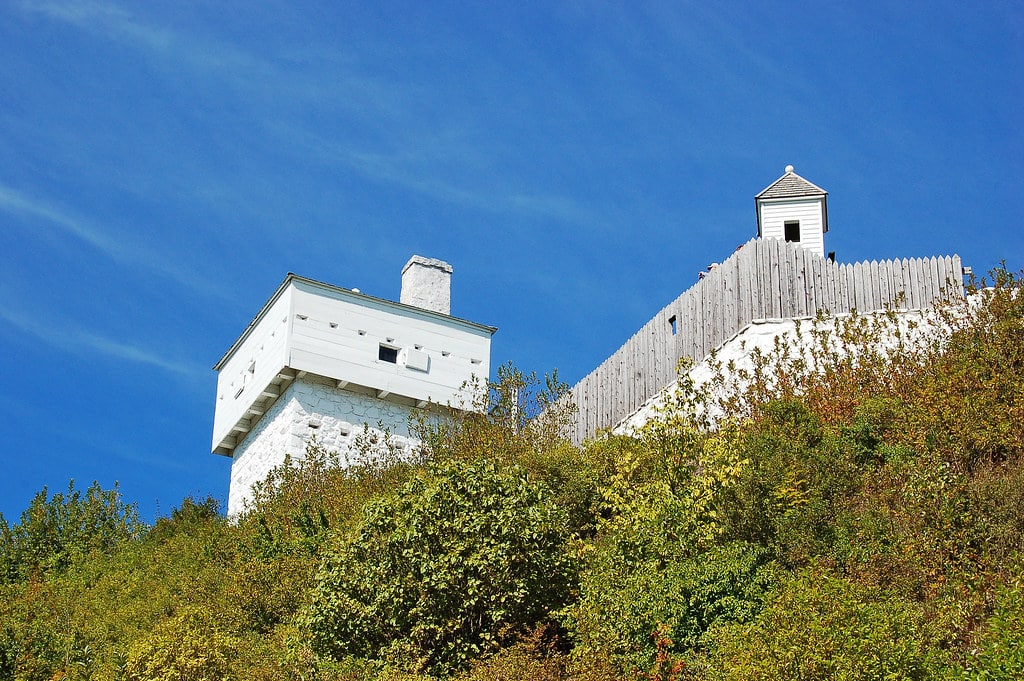A historic white stone fort with a wooden fence and watchtower sits atop a green, shrub-covered hill under a clear blue sky.