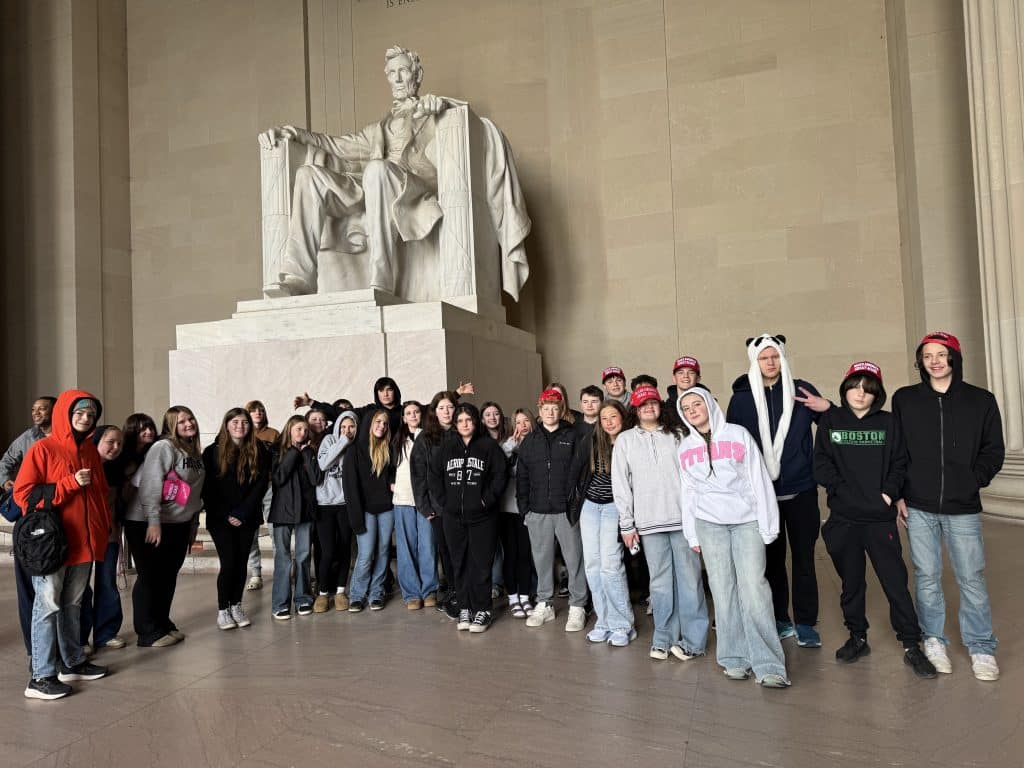 A group of young people pose for a photo in front of the large seated Abraham Lincoln statue at the Lincoln Memorial in Washington, D.C. Many wear casual clothes and hoodies, some with animal ears or hats.