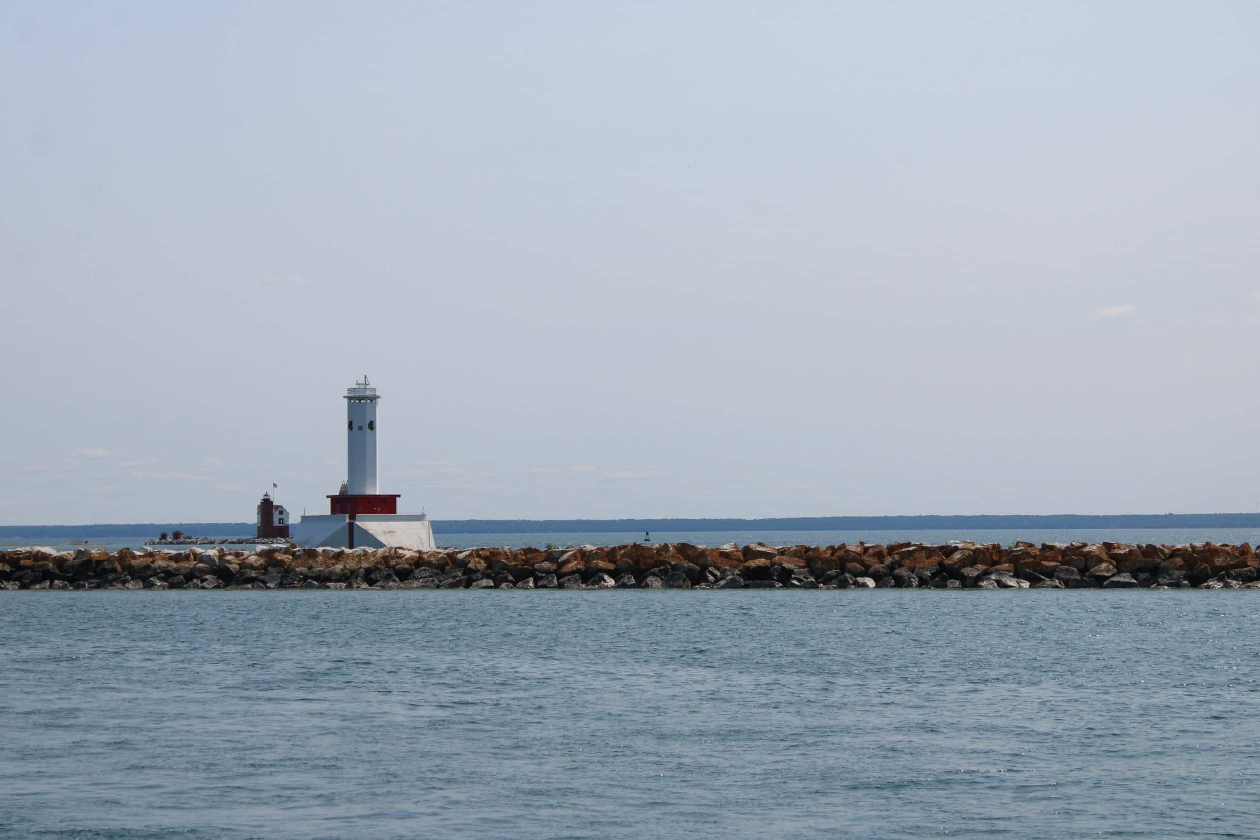 A white lighthouse with a red base stands on a rocky breakwater surrounded by calm blue water under a clear sky. The horizon and distant land are visible in the background.