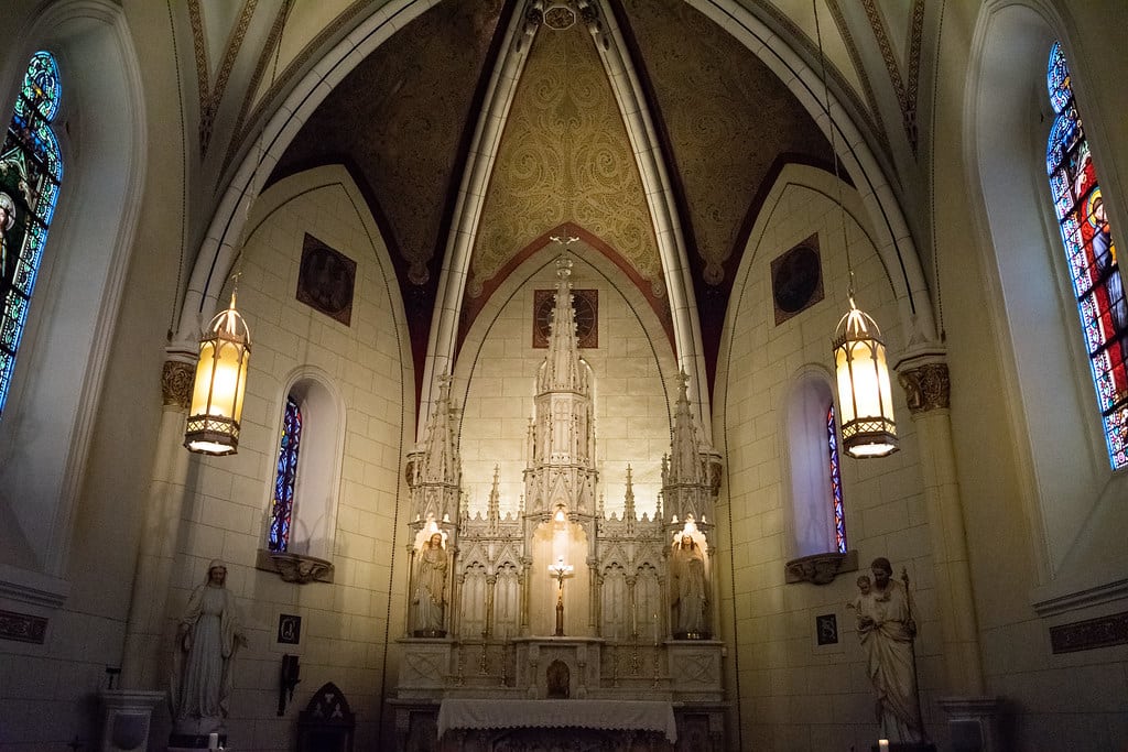 A church altar with intricate white carvings, tall arched ceiling, hanging lanterns, and stained glass windows on both sides—a peaceful spot often listed among the Top 10 things to do on a Santa Fe School Trip. Statues line the walls and soft lighting sets the mood.
