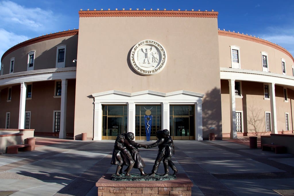 The photo shows the front entrance of the New Mexico State Capitol, one of the Top 10 things to do on a Santa Fe School Trip, with a round emblem above the doors and a bronze statue of children playing in the foreground.