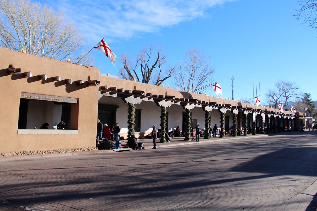 People sit beneath a covered walkway of an adobe building with wooden beams, displaying goods on the ground—an authentic scene and one of the Top 10 things to do on a Santa Fe School Trip. Red and white flags fly above under a clear blue sky.