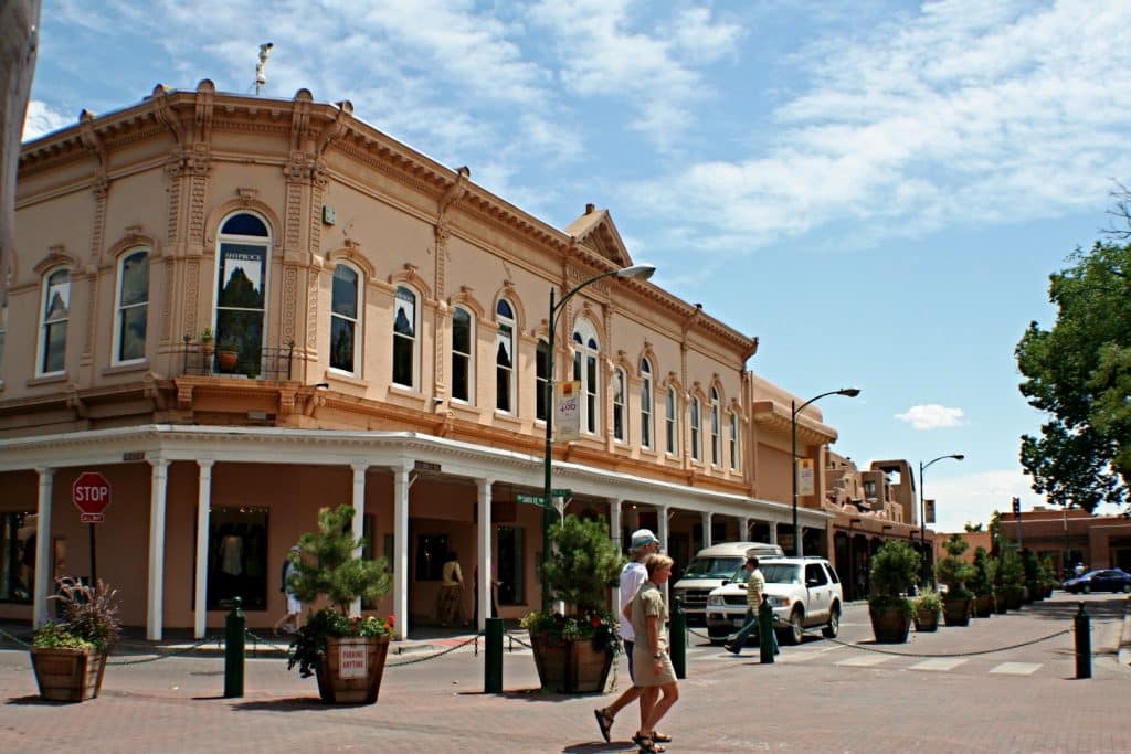 A historic, tan-colored building with arched windows lines a sunny street—perfect for exploring as part of the Top 10 things to do on a Santa Fe School Trip. Two people stroll near potted plants, with parked cars and a stop sign in view under partly cloudy skies.