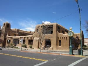 A large adobe-style building with rounded corners and wooden beams stands on a street corner, accompanied by a vintage street clock and clear blue sky—capturing the charm of a Field Trip New Mexico adventure.