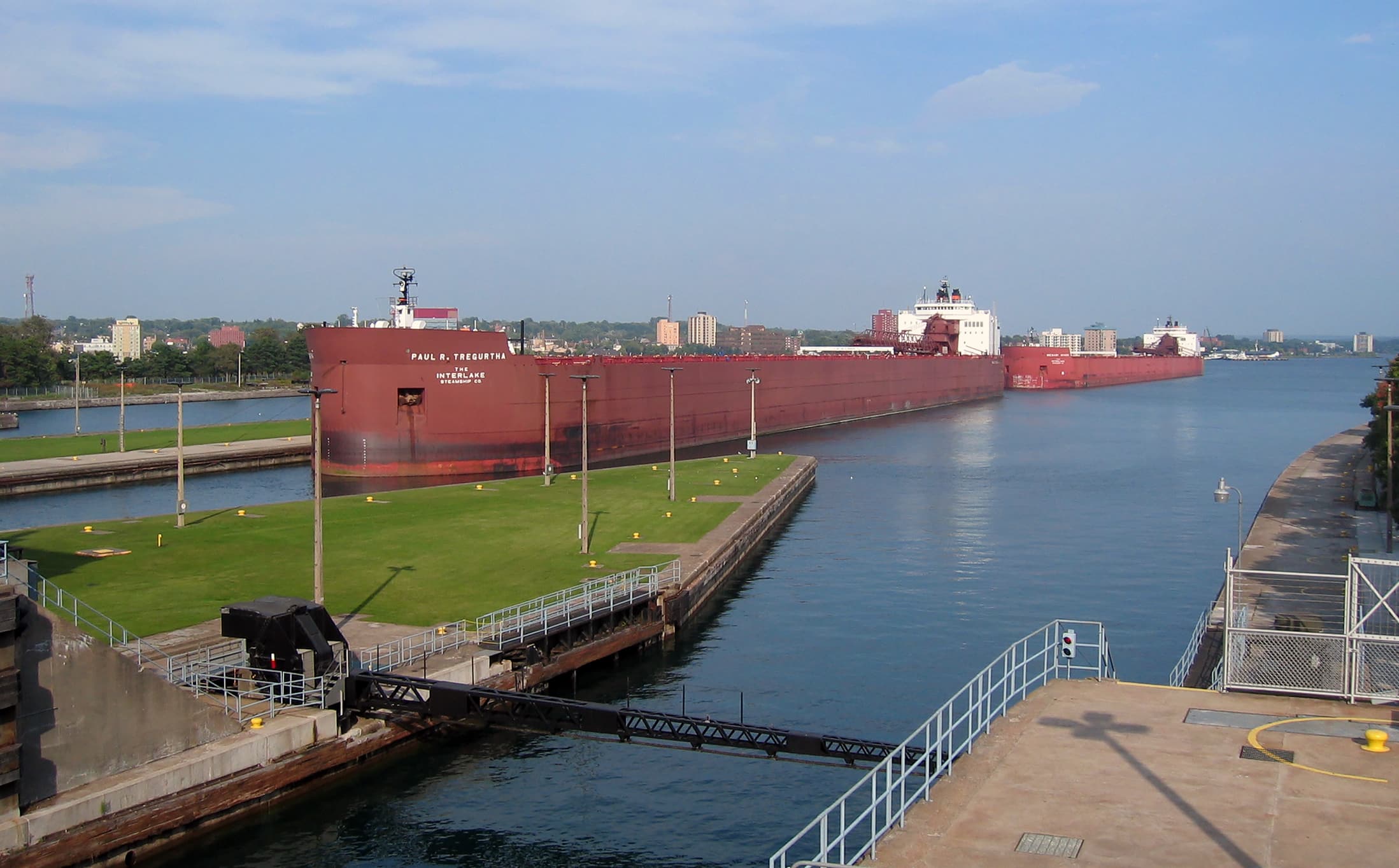A large red cargo ship travels through a canal with grassy areas and buildings on either side; another similar ship follows behind under a blue sky.