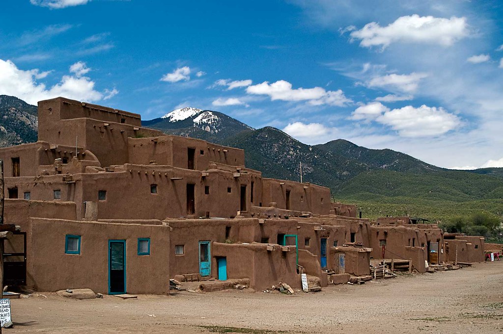 Multi-story adobe buildings with blue doors and windows stand against green hills and a snow-capped mountain—an iconic Field Trip New Mexico scene under a partly cloudy sky.