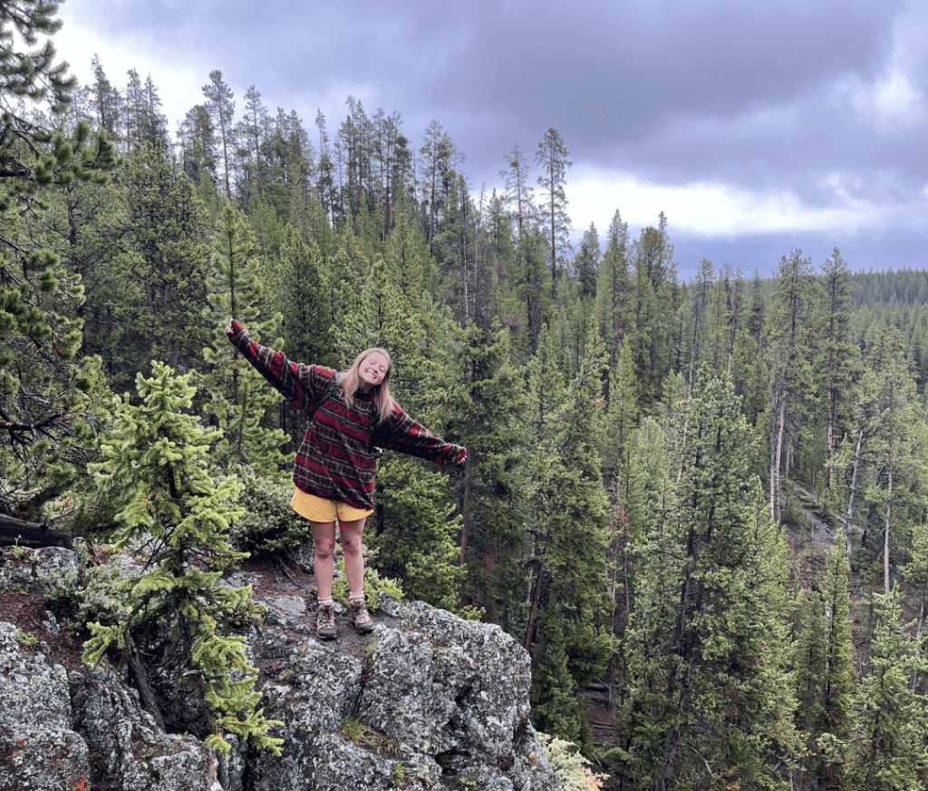 A person stands on a rocky ledge with arms outstretched, smiling, surrounded by dense pine forest under a cloudy sky during a Yellowstone National Park school trip.