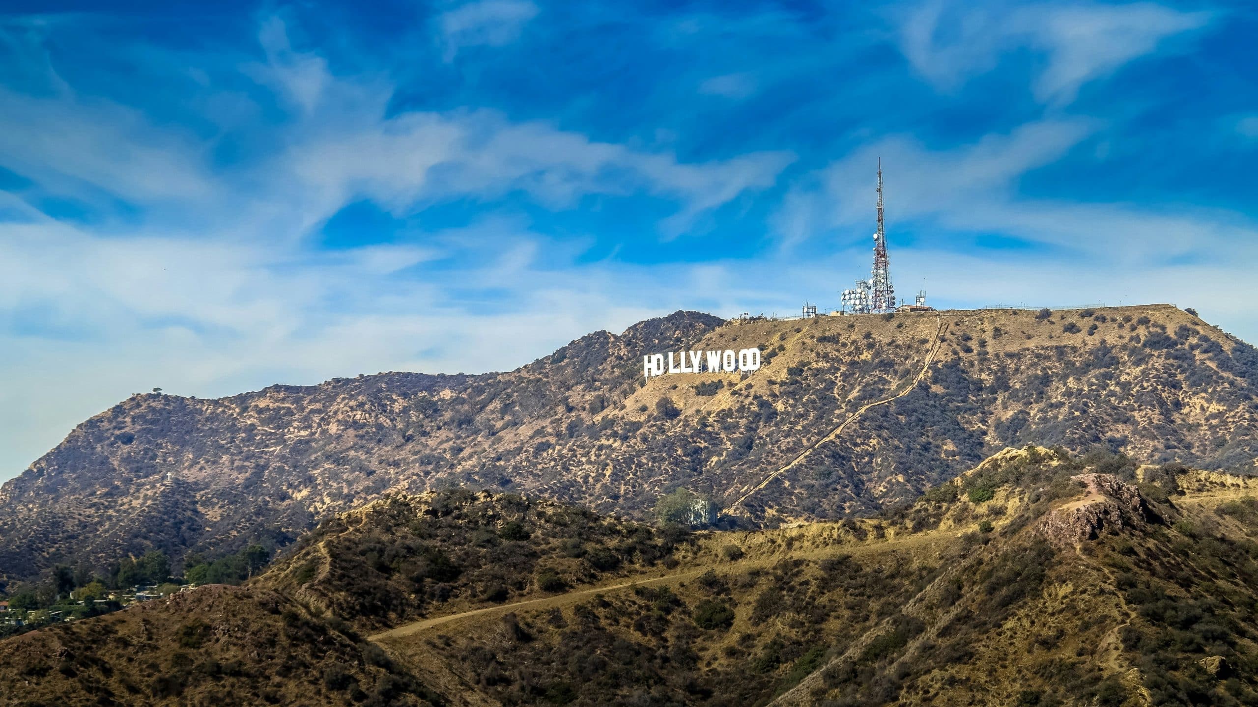 The Hollywood sign sits atop a dry, hilly landscape under a bright blue sky with scattered clouds, making it a classic destination for Los Angeles field trips, with a tall radio tower nearby.