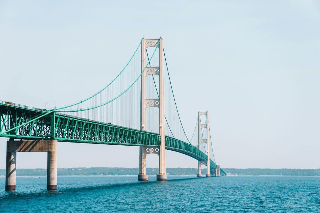 A long suspension bridge with tall towers and green supports spans across blue water, connecting two land masses under a clear sky—a perfect sight during a school trip to Mackinaw City.