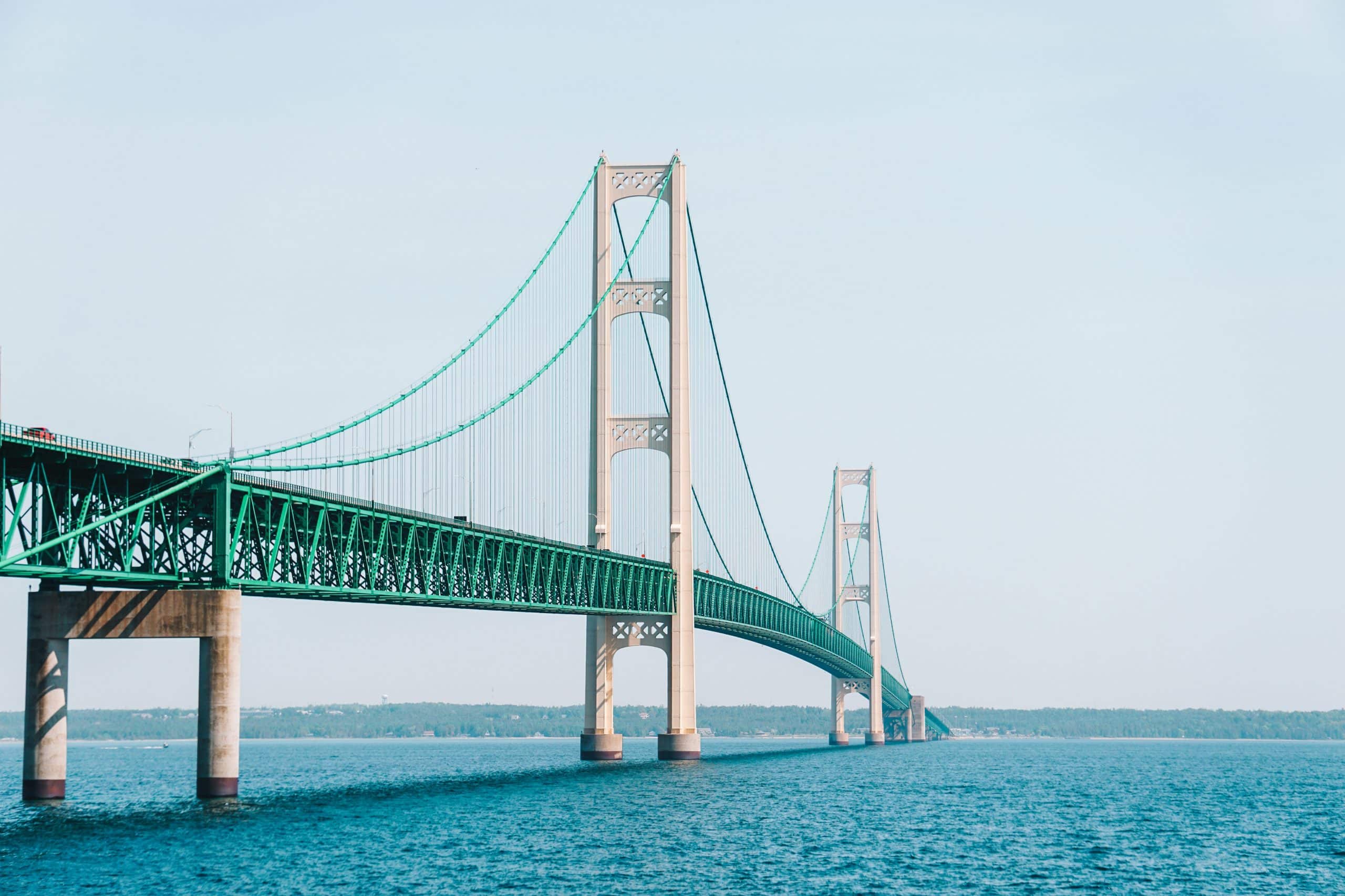 A long suspension bridge with tall towers and green supports spans across blue water, connecting two land masses under a clear sky—a perfect sight during a school trip to Mackinaw City.