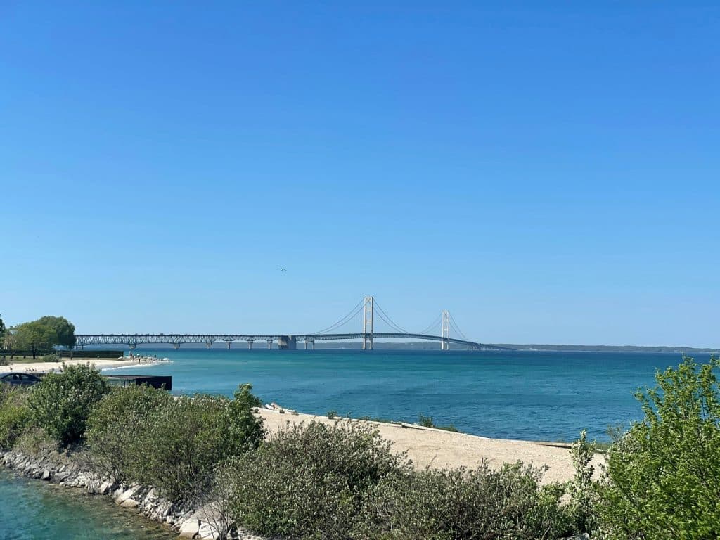 A long suspension bridge spans across blue water under a clear, sunny sky, with green bushes and sandy shorelines in the foreground—a perfect scene for a memorable school trip to Mackinaw City.