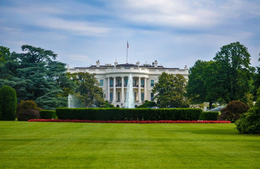 On a School Field Trip from Quaboag MA, students admire the White House, a large white neoclassical building behind manicured lawns and gardens with fountains, surrounded by green trees under a partly cloudy sky.