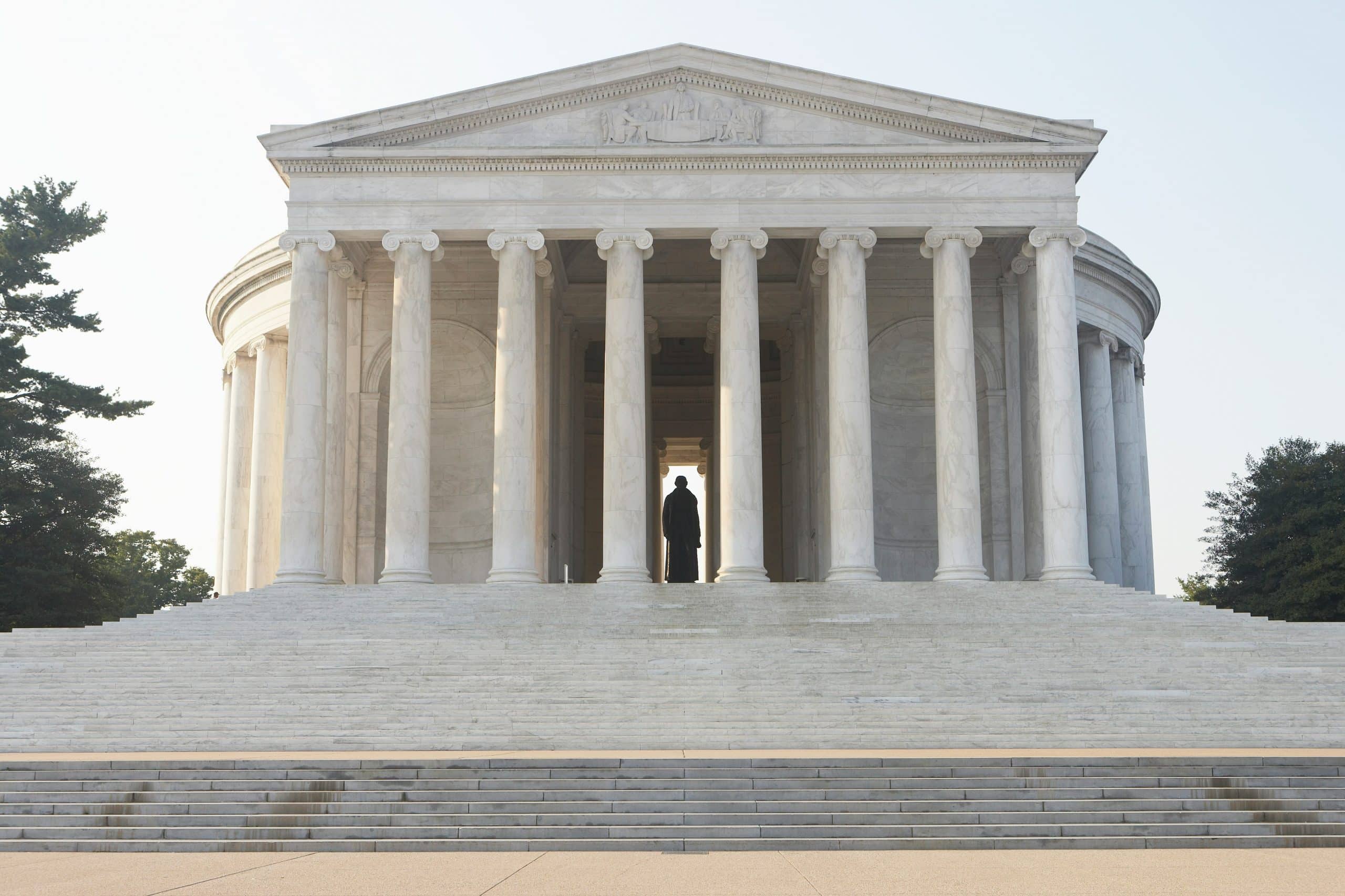 The image shows the Jefferson Memorial in Washington, D.C., with its white marble columns, wide steps, and a statue visible inside the domed structure on a clear day.