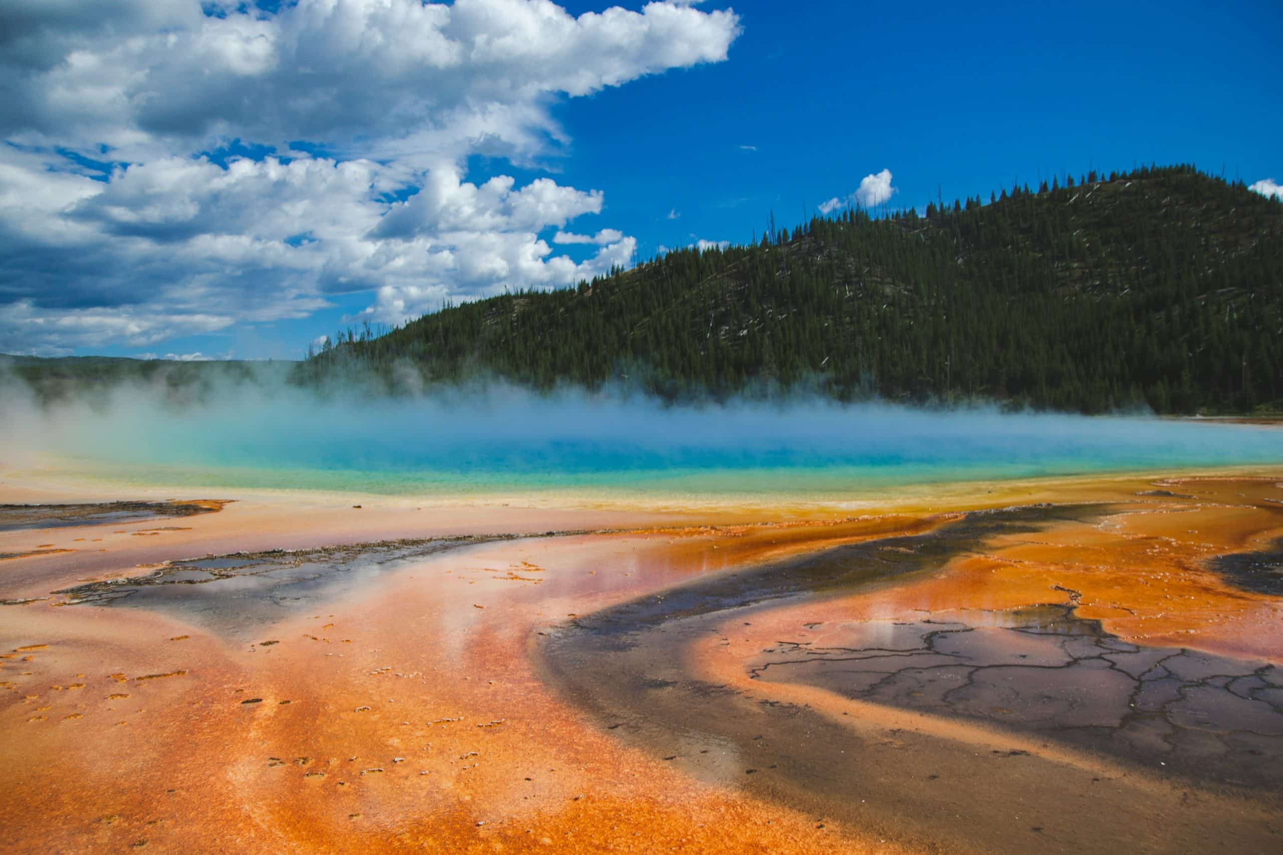 Steaming hot spring with vibrant orange, yellow, and blue colors under a bright blue sky with white clouds in Yellowstone National Park; forested hills in the background.