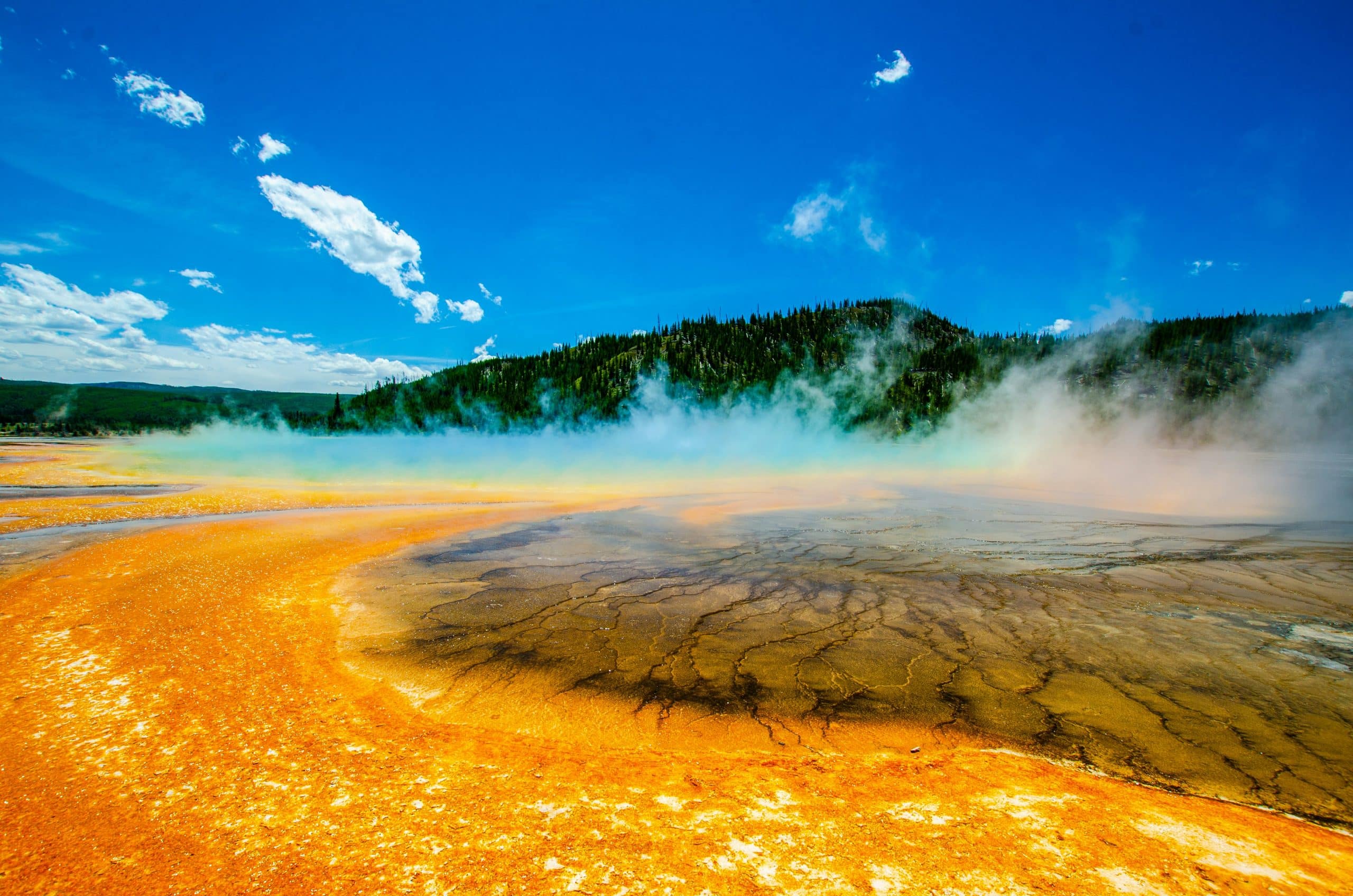 A vibrant hot spring, themed with bright orange and yellow mineral deposits, surrounds steaming, multicolored water, set against a green forest and blue sky with scattered clouds.