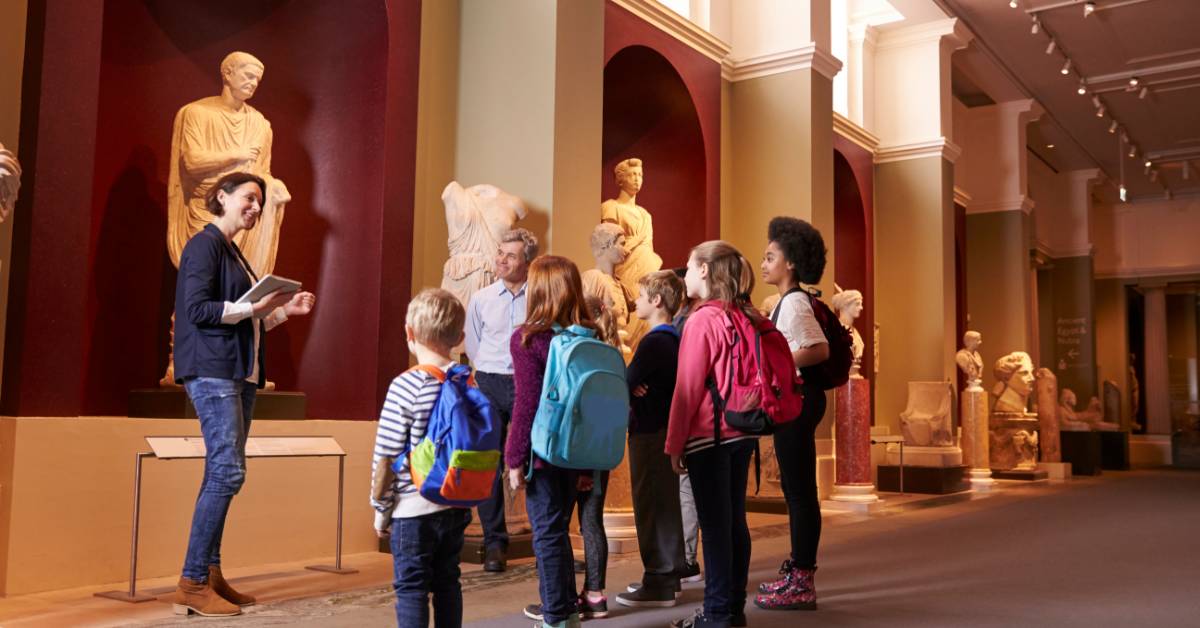 A group of elementary students gathered in front of a statue on a field trip. A teacher stands in front of them to present.