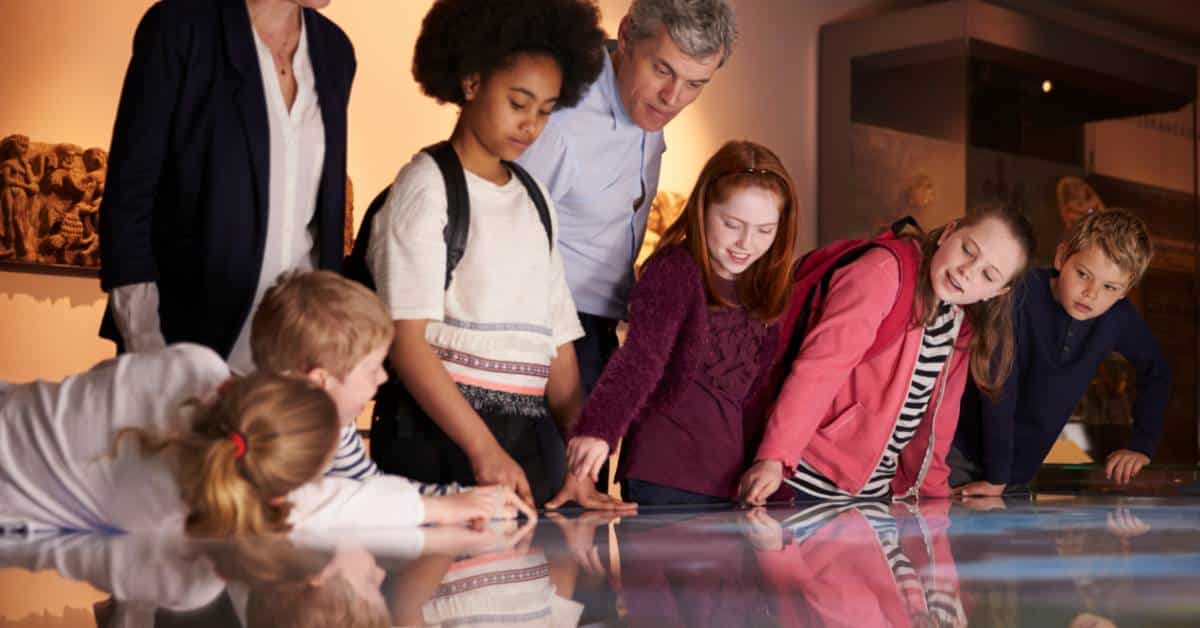 A group of junior high students and two adult chaperones gathered around an exhibit at the museum. They're on a field trip.