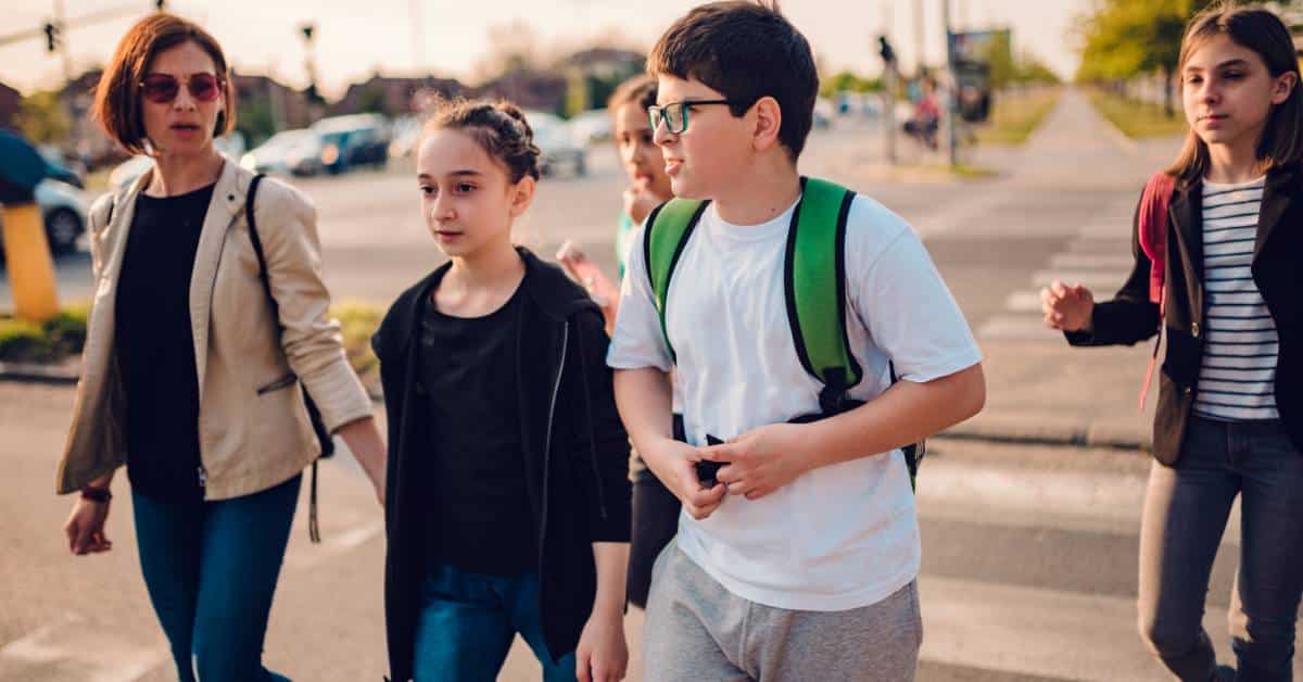 A small group of junior high students crossing the road with their backpacks on. They have a chaperone with them.