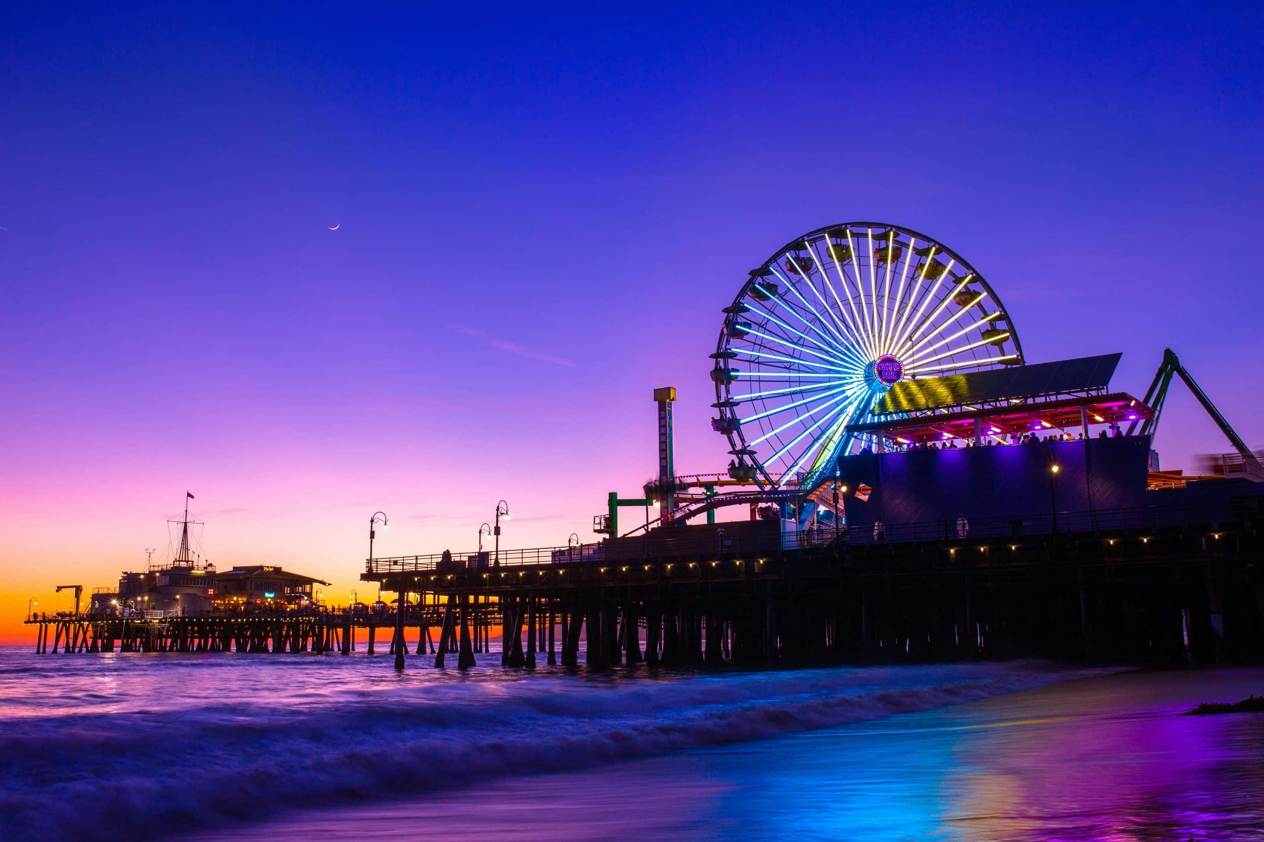 A brightly lit Ferris wheel and amusement park sit on a pier over the ocean at sunset—an iconic spot for Los Angeles field trips, with vivid purple, pink, and blue skies and gentle waves lapping the shore.
