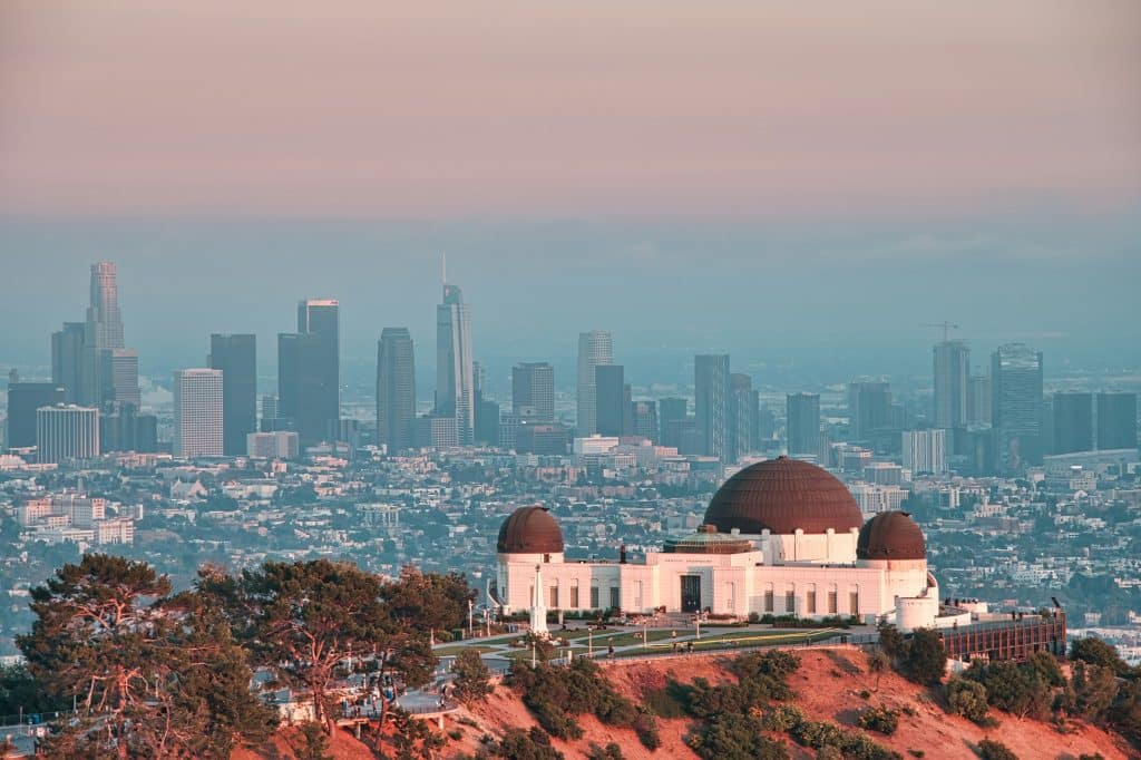 Griffith Observatory sits on a hill with the Los Angeles city skyline, including tall skyscrapers, visible in the background under a hazy sunset sky. Trees frame the foreground, making it a favorite destination for Los Angeles field trips.