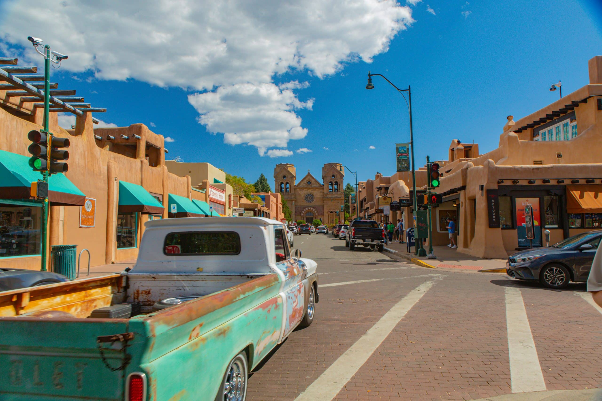 A vintage turquoise pickup truck drives down a street lined with adobe-style buildings under a bright blue sky—one of the top 10 things to do on a Santa Fe school trip is exploring these historic landmarks with your group.