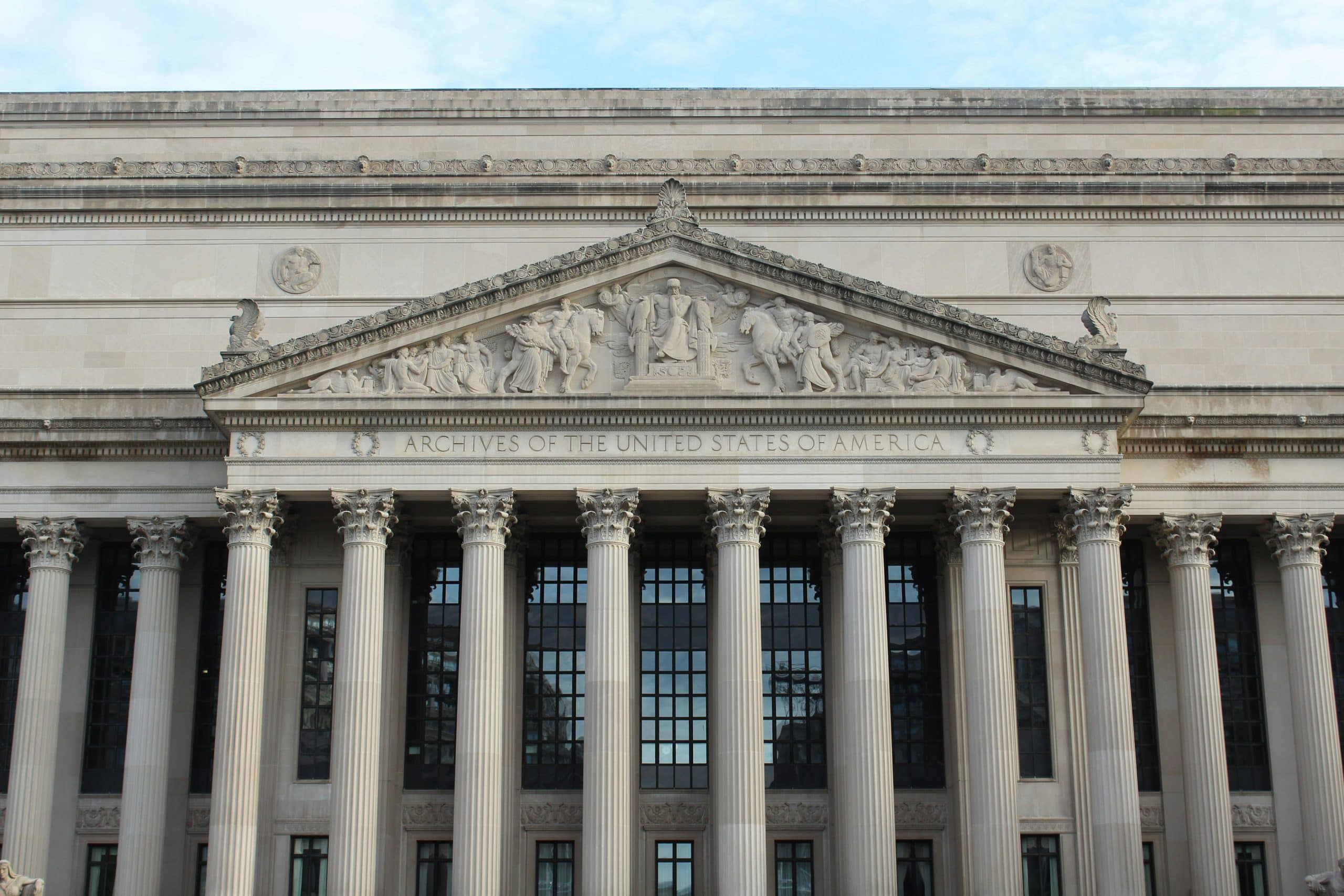 The front facade of the U.S. National Archives building, featuring tall columns, a detailed pediment with sculptures, and the inscription “ARCHIVES OF THE UNITED STATES OF AMERICA.”.