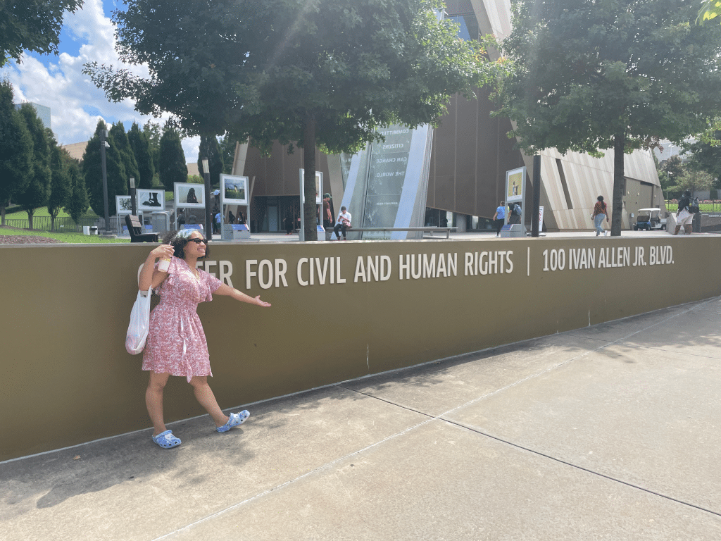 A woman in a pink dress and blue shoes points to the sign for the Center for Civil and Human Rights, a landmark site perfect for Educational Tours Registration, located at 100 Ivan Allen Jr. Blvd on a sunny day outside a modern building.