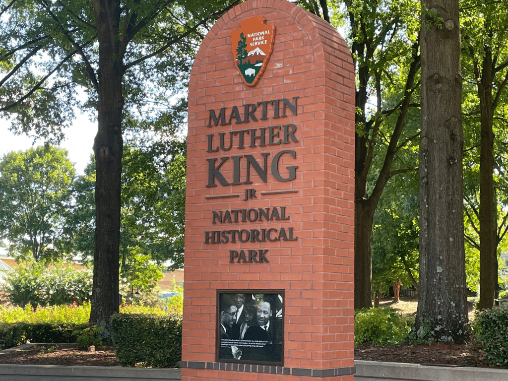 A brick sign reads "Martin Luther King Jr. National Historical Park" with the National Park Service emblem on top. Perfect for Landmark Educational Tours, the site features trees and greenery, plus a photo of Dr. King at the bottom of the sign.