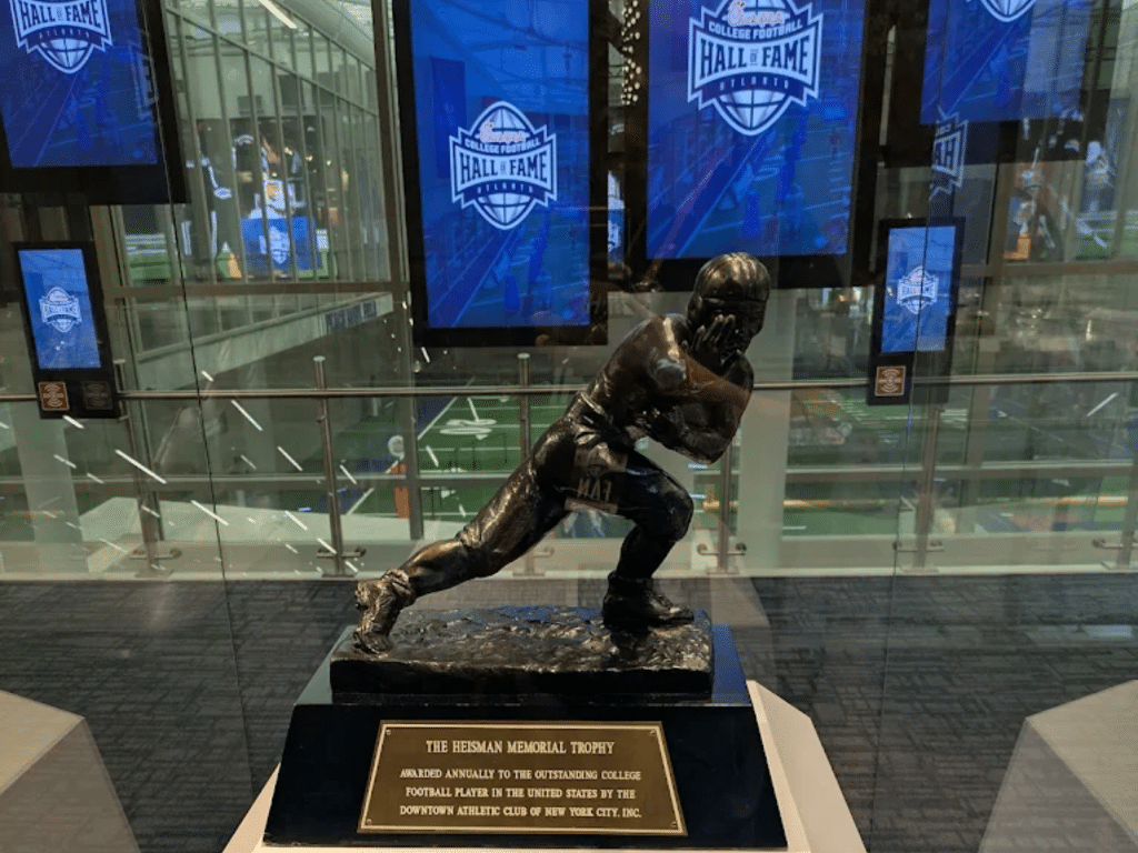 A bronze Heisman Trophy statue is displayed in a glass case, with blue Hall of Fame screens and a football stadium visible in the background. The plaque explains its significance for guests on Landmark Educational Tours Registration.