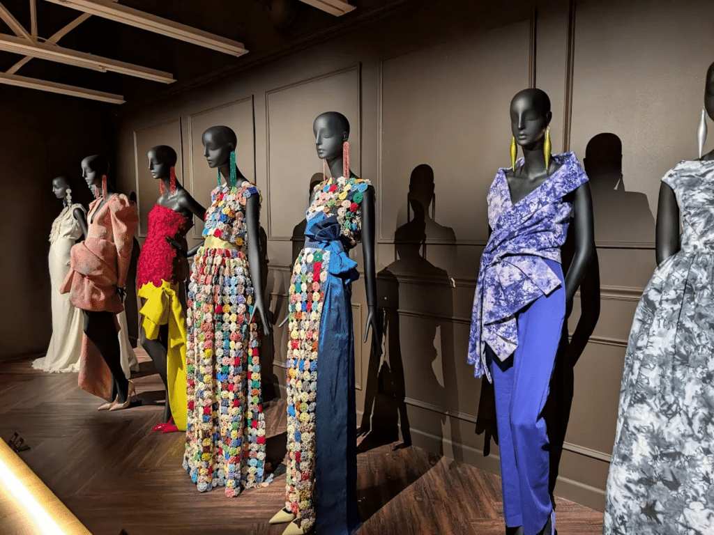 Mannequins dressed in colorful, elaborate dresses with floral patterns and bold textures stand in a row against a dark wall at the Landmark Educational Tours Registration fashion exhibit.