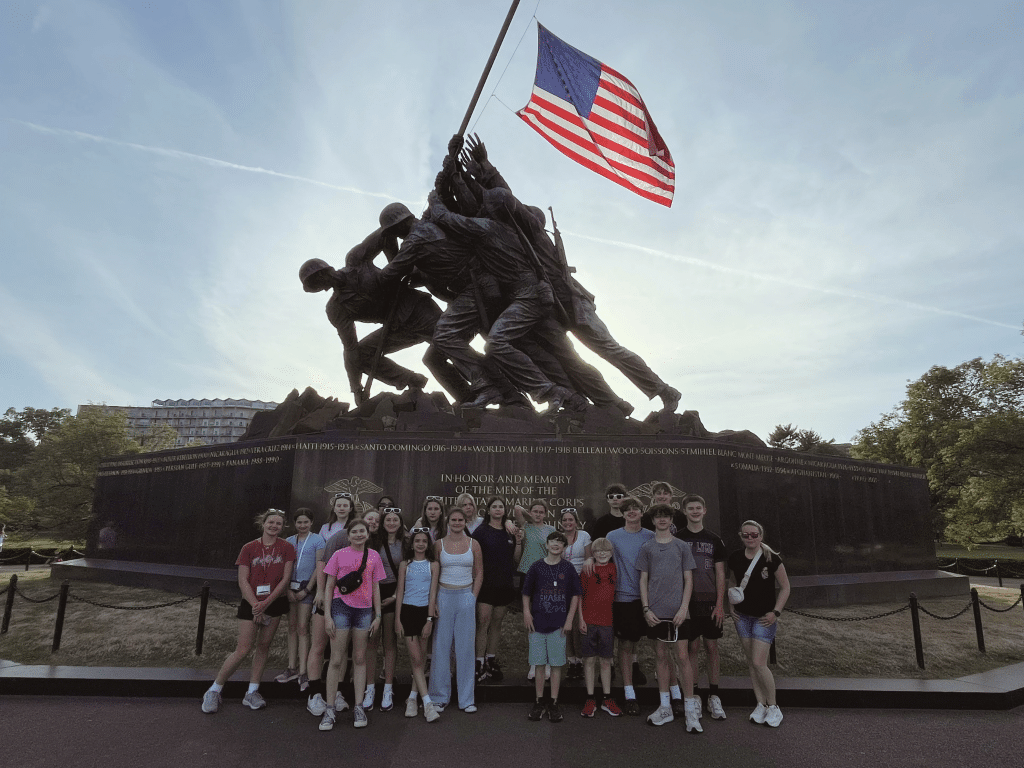 A group of people poses in front of the U.S. Marine Corps War Memorial, featuring soldiers raising an American flag. The sky is clear and the sun shines from behind the statue.