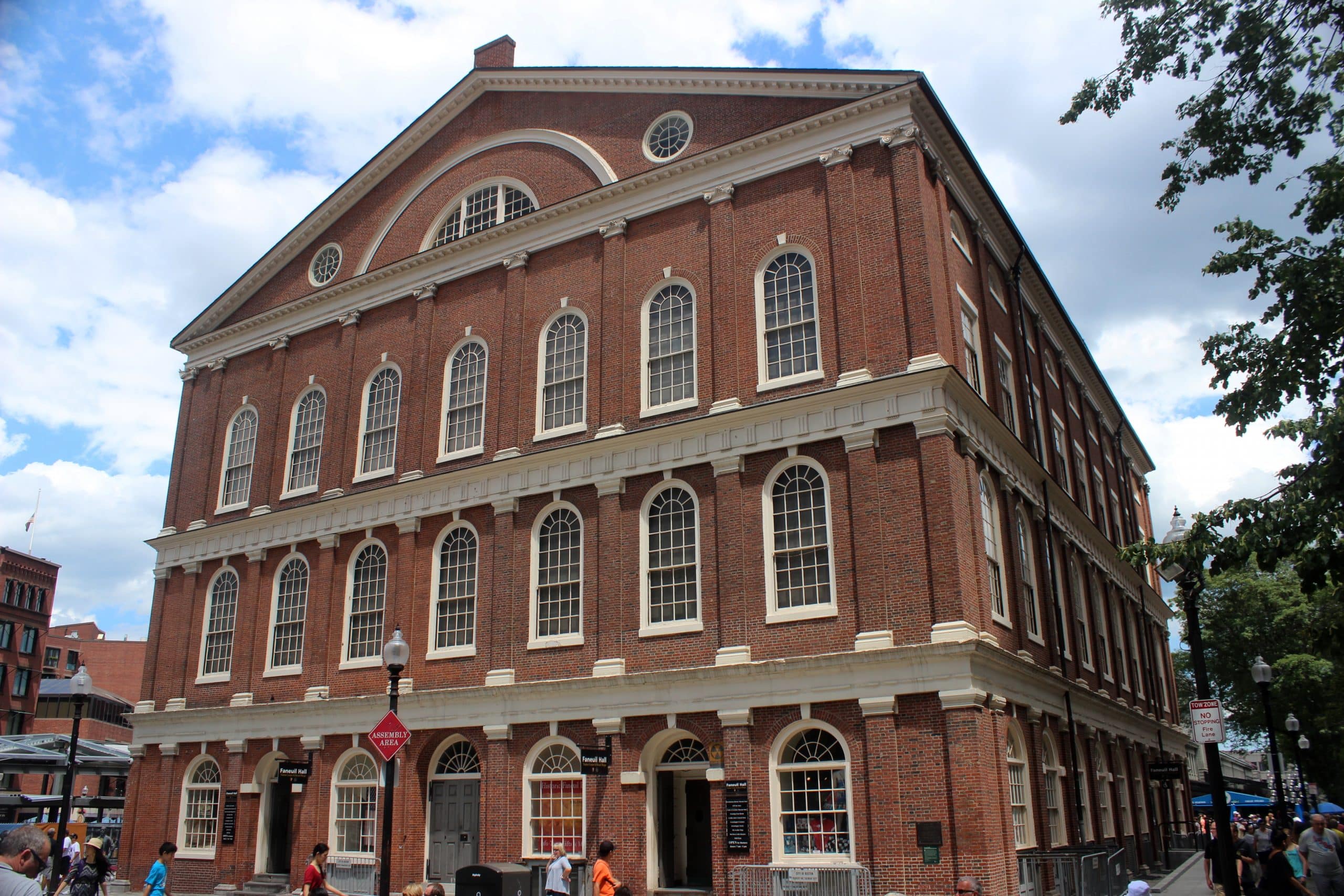 A large historic brick building with three stories and many tall arched windows; people walk nearby under a partly cloudy sky.