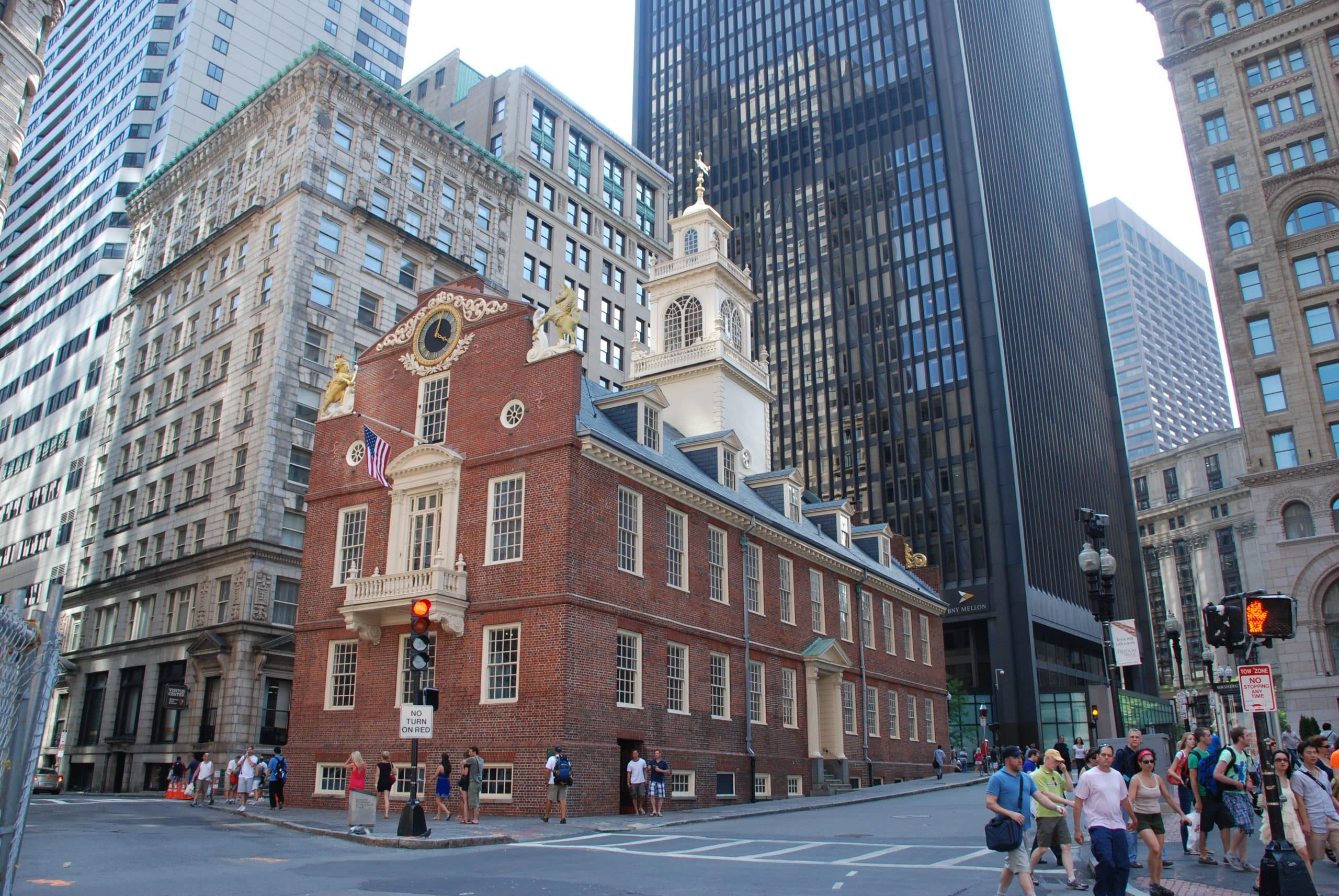 A historic red-brick building with a white steeple stands at a city intersection, surrounded by modern and older high-rise buildings—a perfect sight to capture during a School Trip to Boston, with people walking and cars waiting at traffic lights.