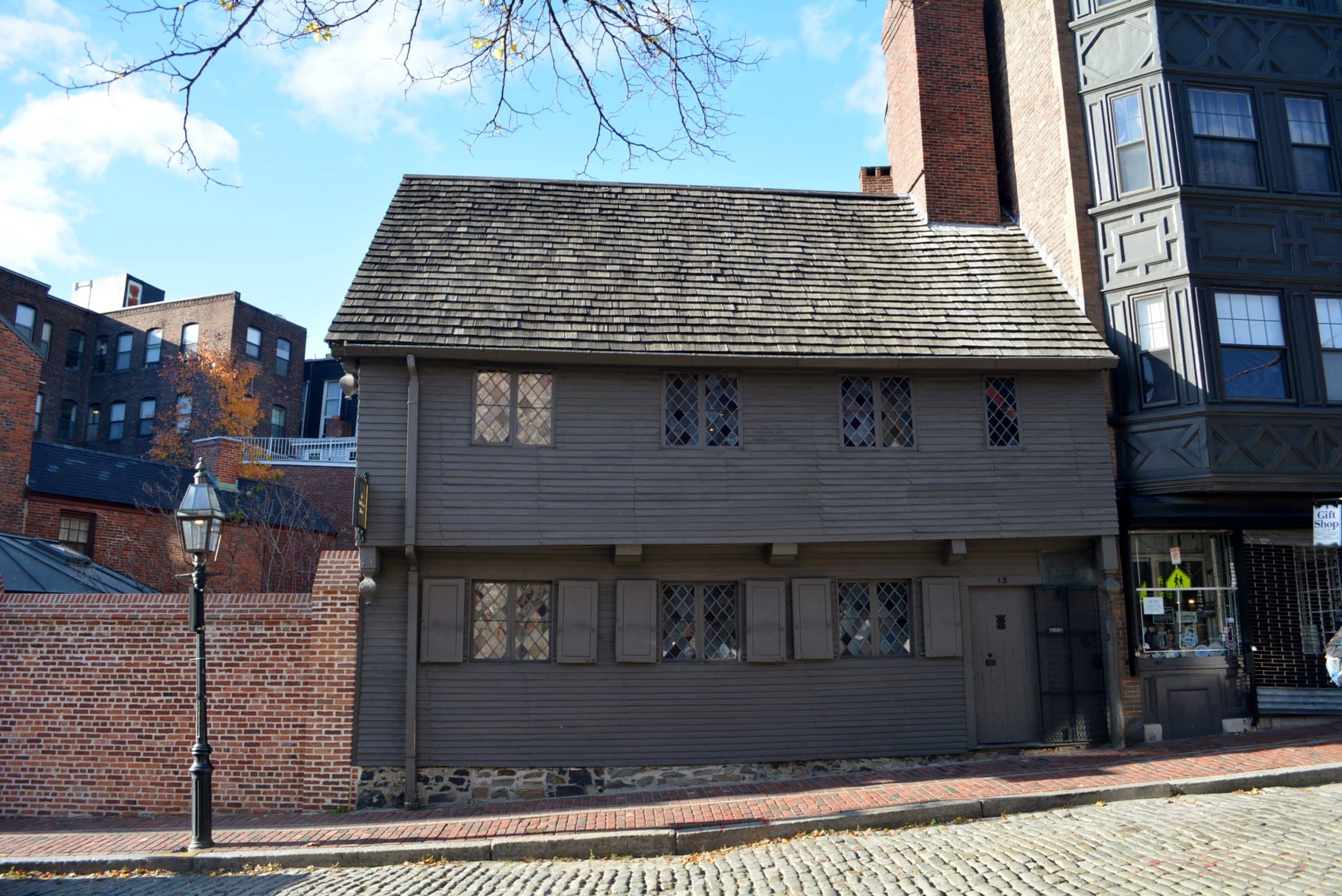 A historic, brown wooden house with diamond-paned windows and a steep roof stands on a cobblestone street—an ideal sight during a School Trip to Boston—bordered by a red brick wall and next to a tall, dark building under a blue sky.