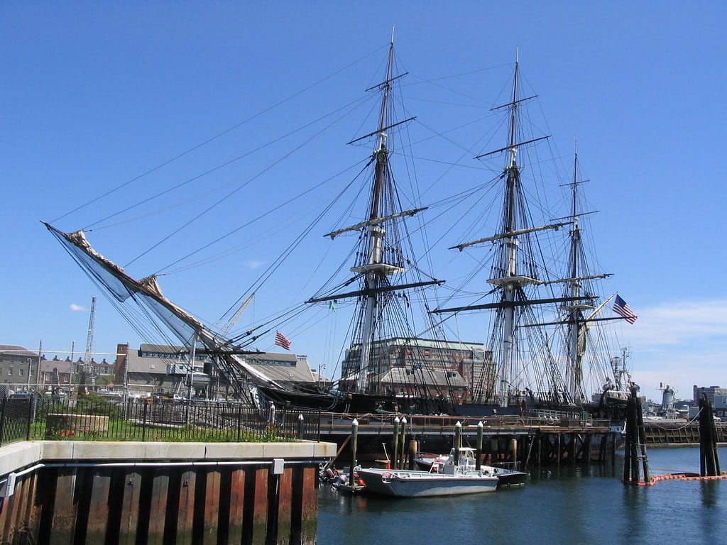 A large historic wooden sailing ship with three tall masts is docked at a harbor, American flags flying, small boats alongside—an iconic scene often admired during a school trip to Boston, with buildings in the background under a clear blue sky.
