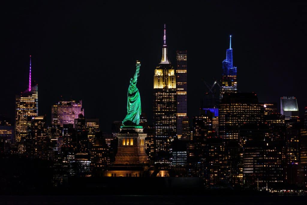 The Statue of Liberty stands illuminated in front of the brightly lit New York City skyline at night, hinting at Student Travel Trends for 2027 as more students seek iconic destinations like NYC with landmarks and stunning city views.