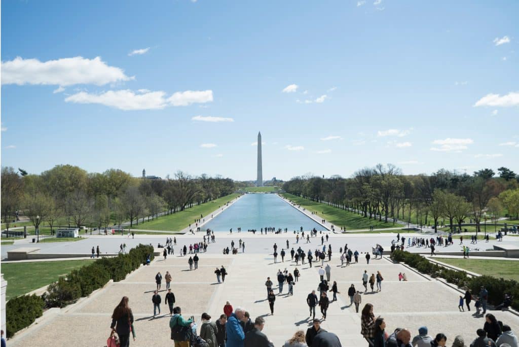 People walk along the National Mall in Washington, D.C., with the Reflecting Pool and Washington Monument visible—an iconic scene expected to feature in student travel trends for 2027 under a blue sky with scattered clouds.