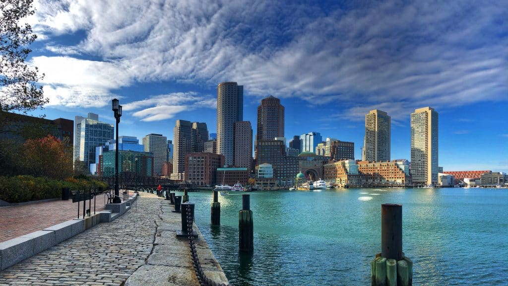 A scenic view from a school trip to Boston—tall skyscrapers, blue sky with dramatic clouds, and calm water bordered by a stone walkway and wooden posts.