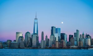 A view of the New York City skyline at dusk, featuring the One World Trade Center and surrounding skyscrapers, with a clear sky and visible moon—perfect for visitors exploring the Best Museums in NYC for Educational Travel. Water shimmers in the foreground.