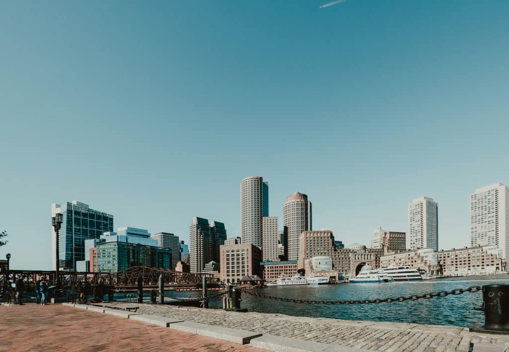 A waterfront view of a city skyline with modern high-rise buildings, a harbor with a docked boat, and a brick walkway in the foreground under a clear blue sky—perfect scenery for your 3 Day Boston Field Trip.