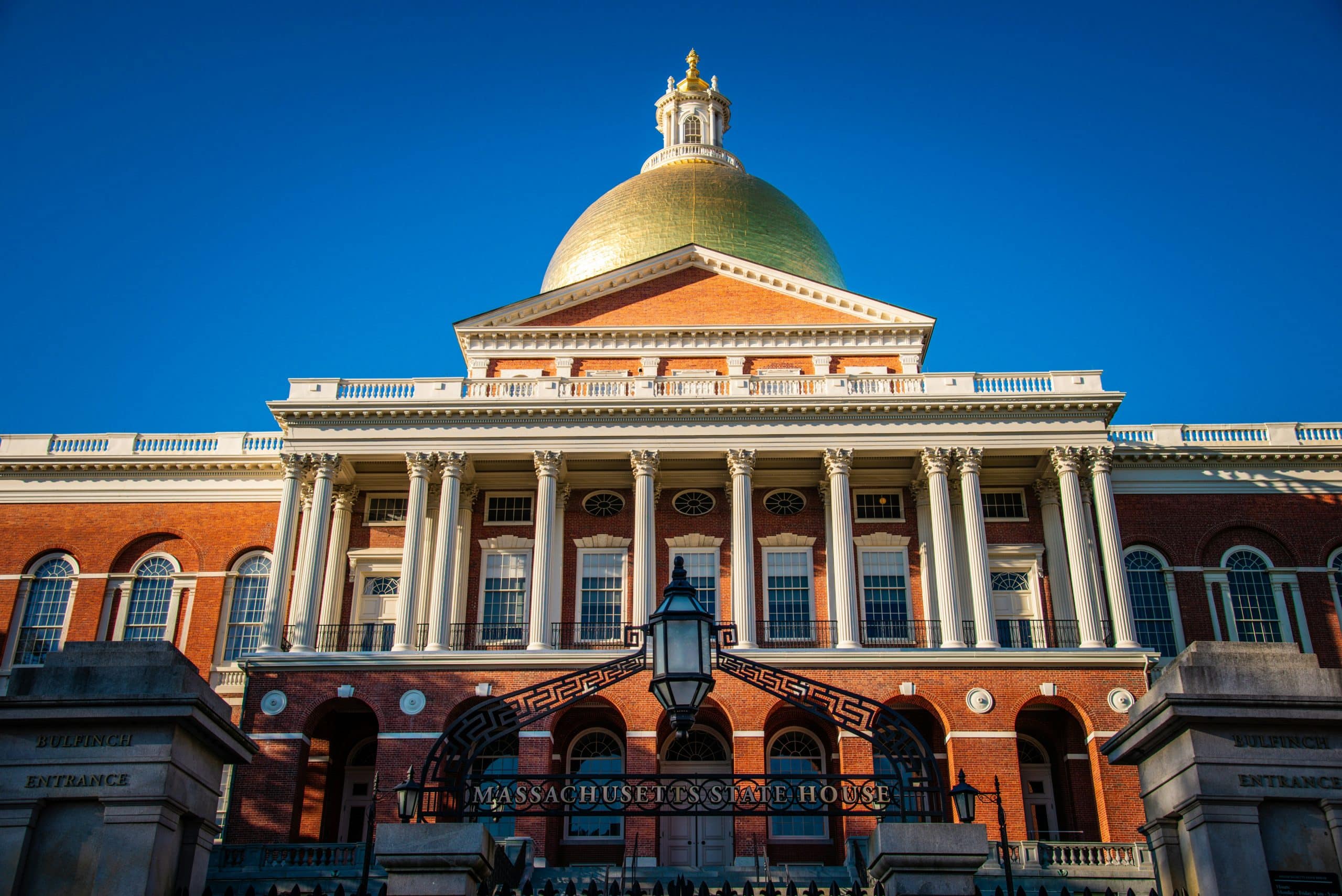 A front view of the Massachusetts State House with its iconic golden dome, white columns, and red brick facade—an essential stop on any 3 Day Boston Field Trip—stands under a clear blue sky.