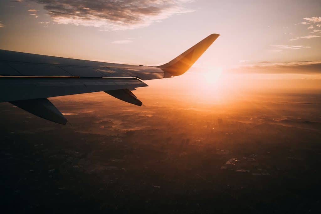 View from an airplane window showing the wing against a golden sunrise or sunset sky, with sunlight illuminating clouds and The Most Popular Student Travel Destinations for 2027 unfolding below.