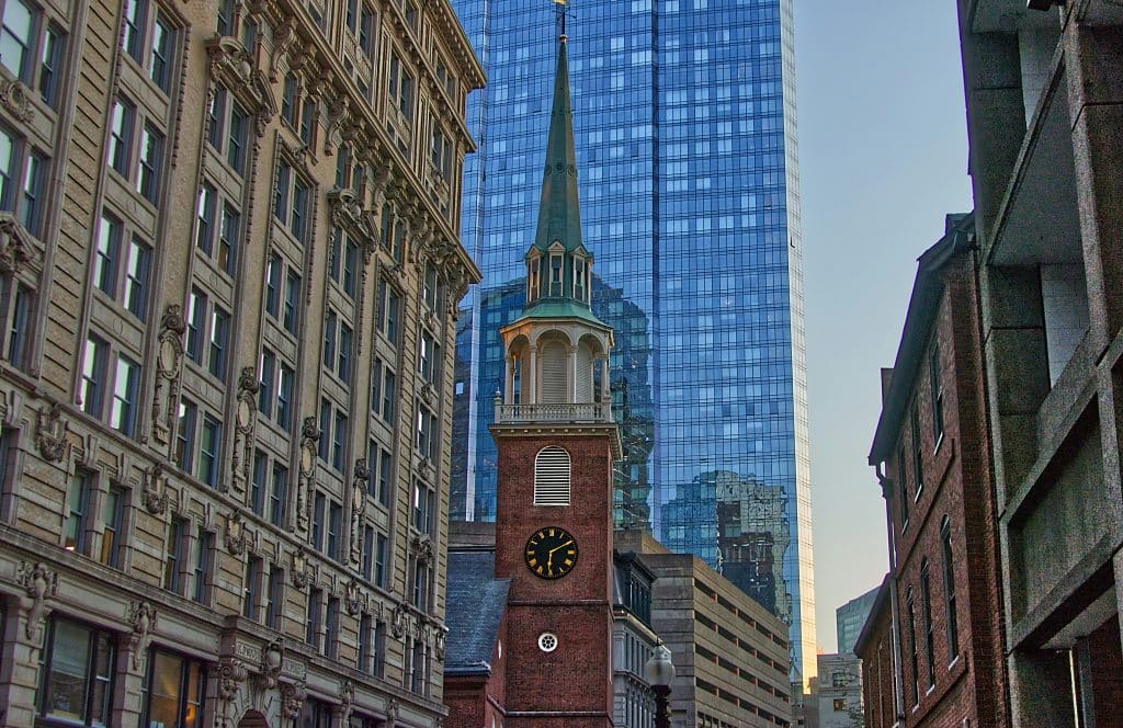 A historic brick church with a tall green steeple and clock stands between older stone buildings, with a modern glass skyscraper rising behind—an iconic view perfect for a School Trip to Boston, all bathed in daylight.
